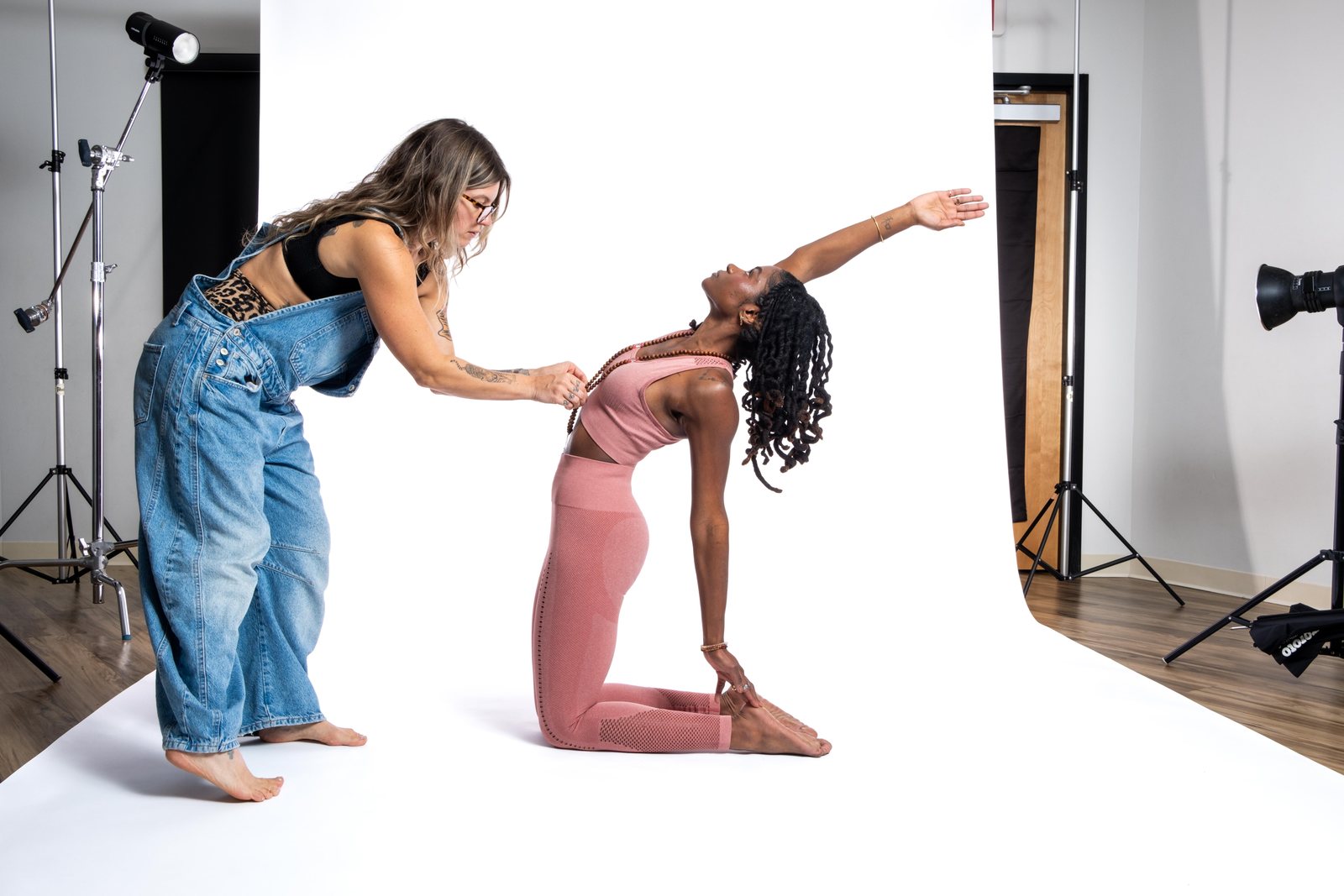 In the studio at Tru Yoga, on a white seamless backdrop between studio lights, a woman in denim overalls adjusts the shoulders of a teacher kneeling in camel pose.