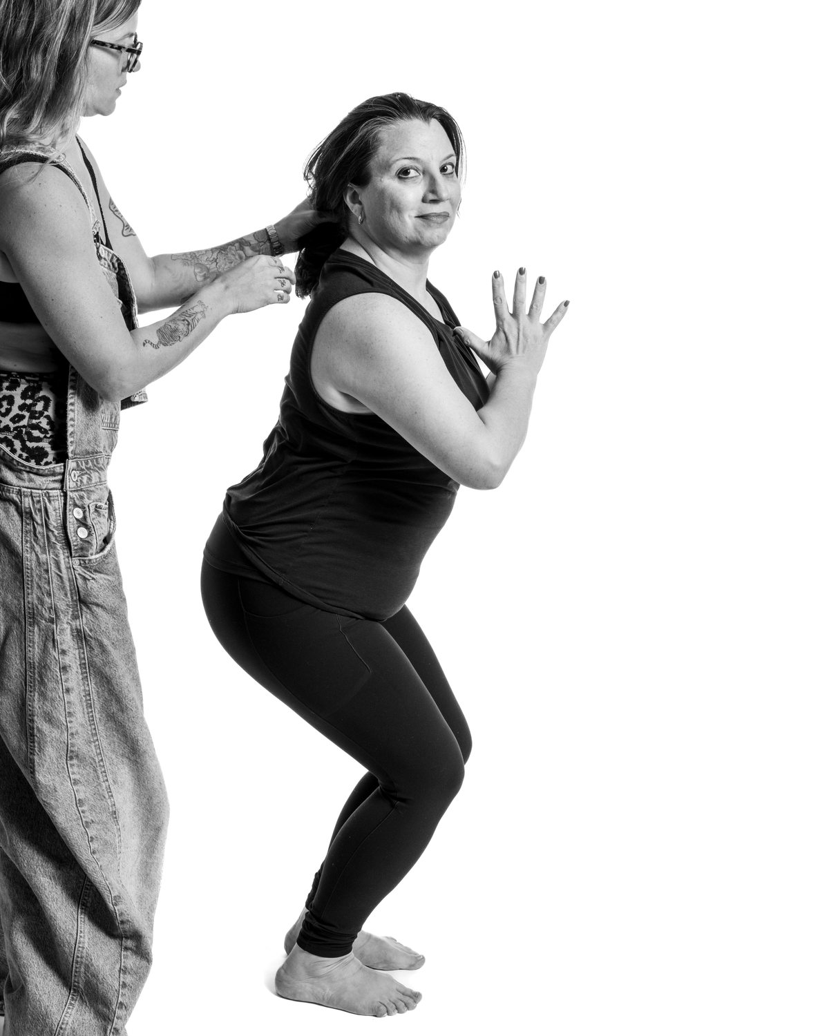 A woman in denim overalls steadies a teacher holding a low squat, hands pressed together at her chest.