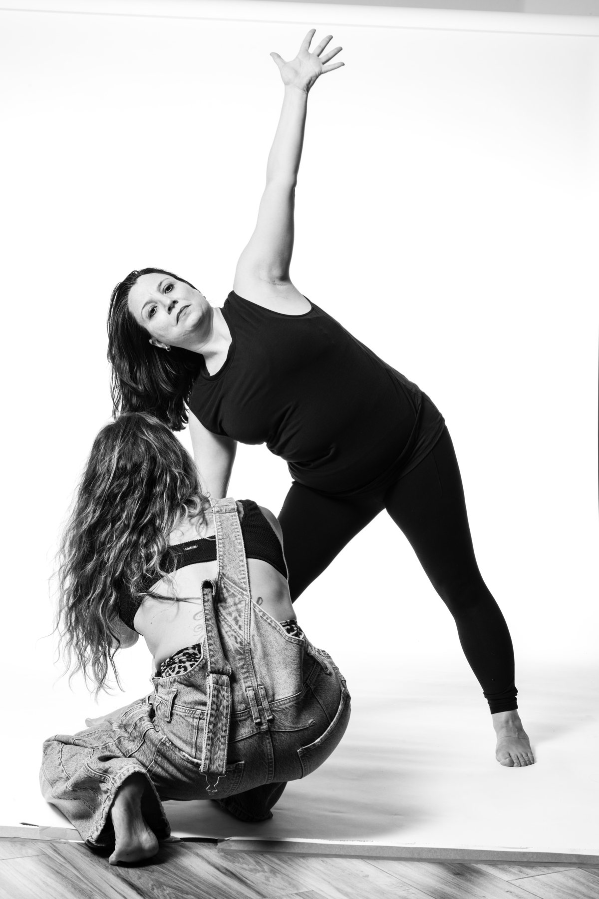 A teacher reaches one arm up in a wide side stretch while a woman in denim crouches beside her.