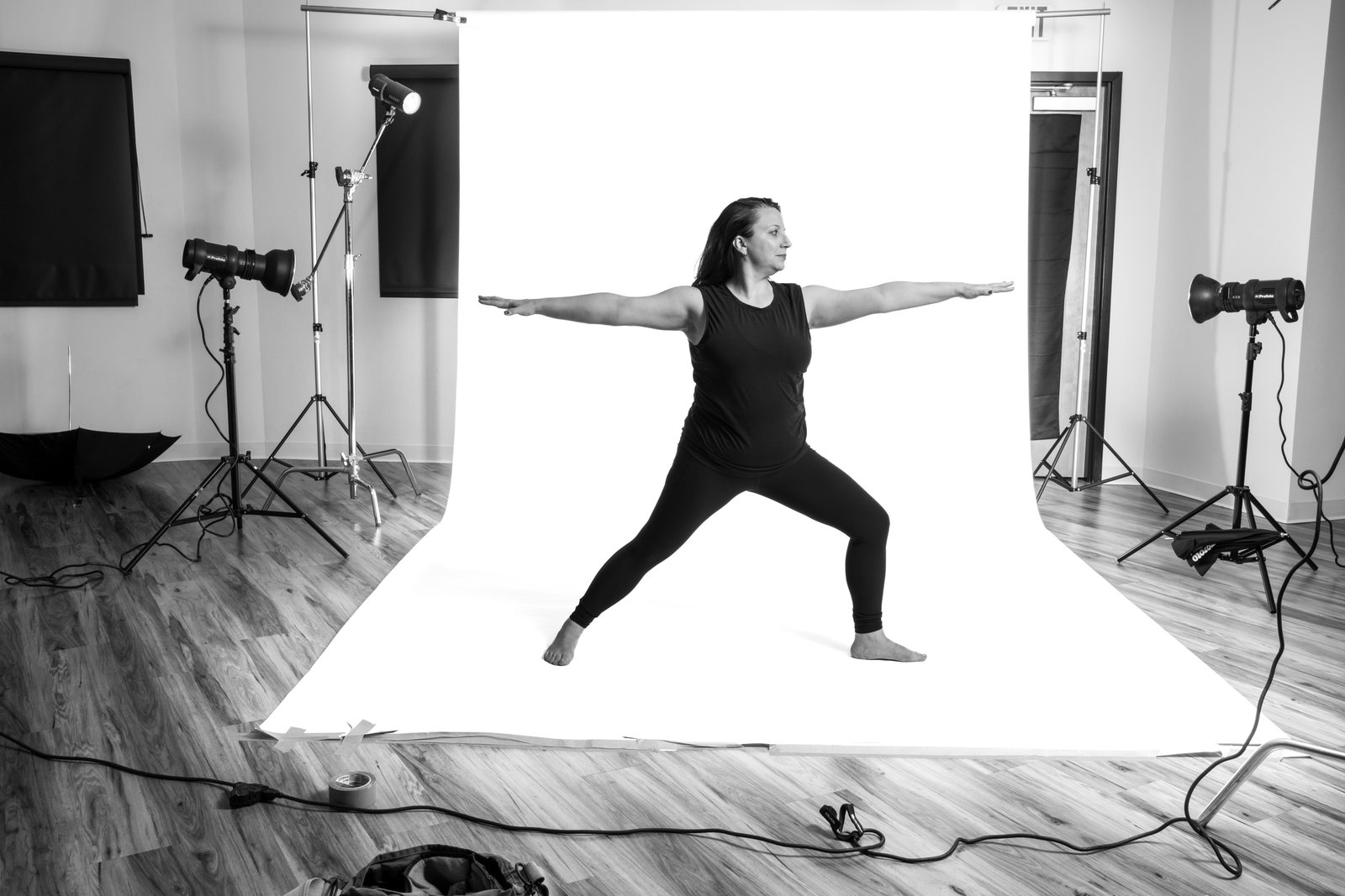 A teacher in warrior pose at the centre of the white seamless, with the whole studio around her: lights, stands, cables and the wooden floor.