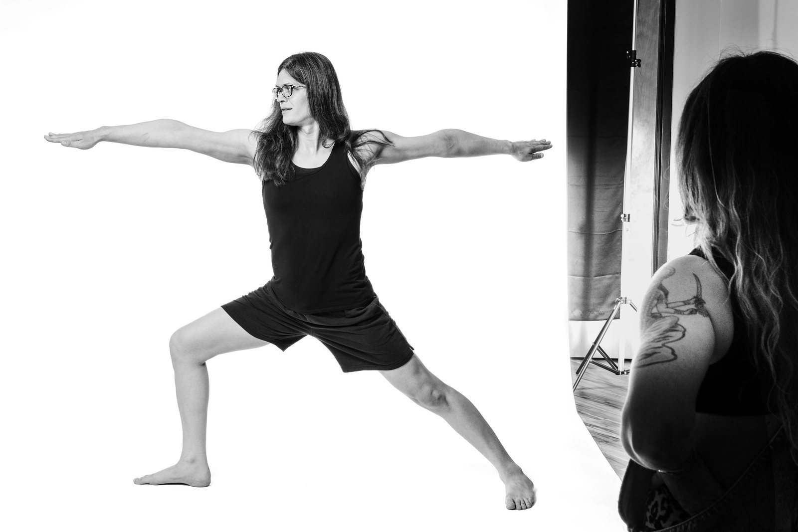 A teacher in glasses holds warrior pose on white while a tattooed figure watches from the right edge of the frame.