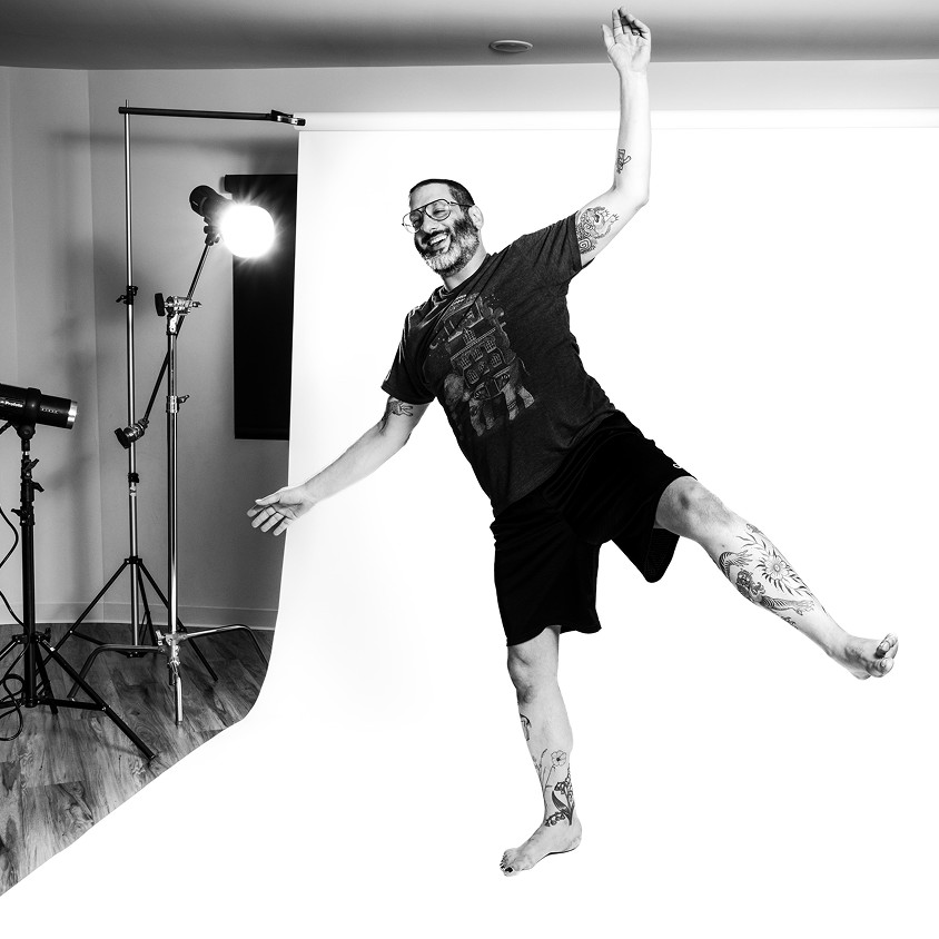 A bearded man in glasses laughs mid-kick on the white seamless, one arm flung up, studio lights behind him.
