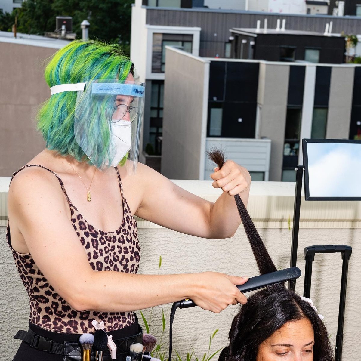 A rooftop portrait session: one person in a mask and face shield working on another person’s hair.